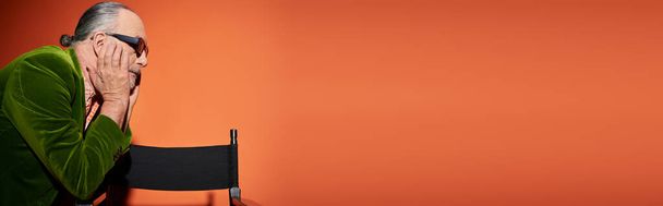vista lateral del modelo masculino senior en chaqueta de terciopelo verde y gafas de sol oscuras de pie cerca de la silla, tocando la cara y pensando en el fondo rojo y naranja, estilo personal, moda casual, pancarta  - Foto, Imagen
