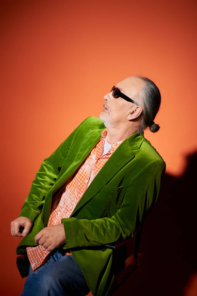 surprised senior man in dark sunglasses, trendy shirt and green velour blazer sitting on chair and looking away on red and orange background with shadow, fashion look, positive aging concept - Foto, afbeelding