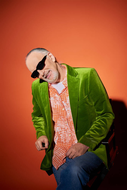 vrolijke senior man met grijs haar en baard zittend op stoel en lachend naar camera op rode en oranje achtergrond, donkere zonnebril, groene velours blazer, modetrend, happy aging concept - Foto, afbeelding