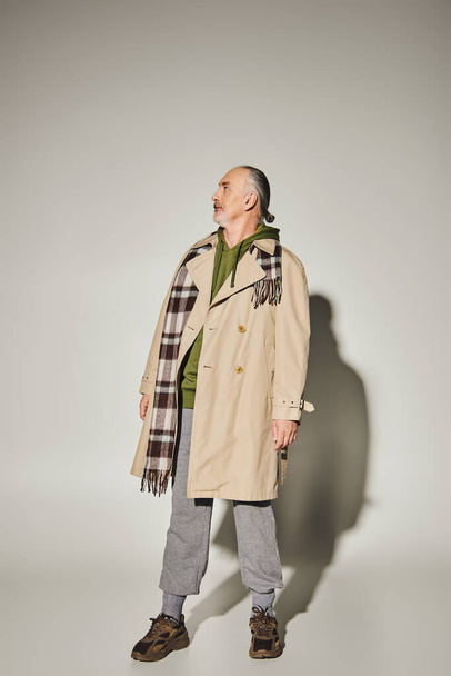 full length of senior male model in beige trench coat, plaid scarf and green hoodie standing on grey background with shadow and looking away, stylish casual attire, fashionable aging concept - Photo, image