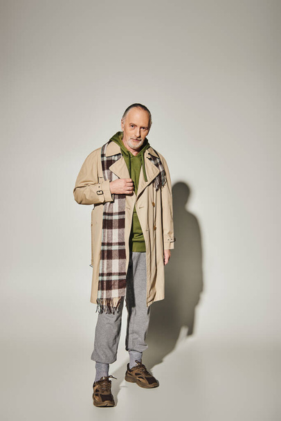 fashion look, positive aging, full length of smiling senior male model in green hoodie, beige trench coat and plaid scarf standing and looking at camera on grey background with shadow - Photo, image