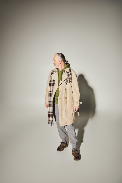 full length of aged and bearded man in beige stylish trench coat, green hoodie and plaid scarf standing and looking away on grey background with shadow and copy space, fashion and age concept - Photo, Image