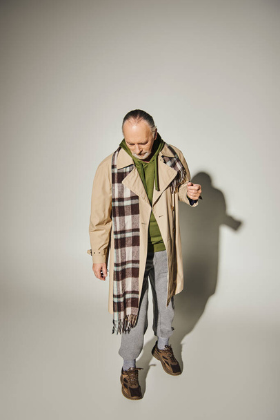 full length of aged bearded man looking down while standing in trendy casual attire on grey background with shadow, beige trench coat, plaid scarf, green hoodie, fashion and age concept  - Foto, imagen