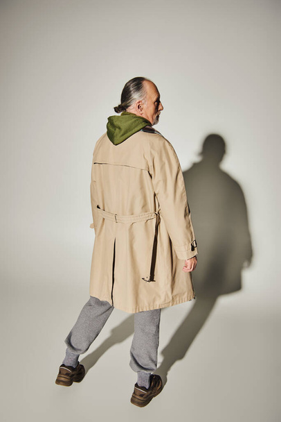 back view of senior model in stylish casual clothes standing on grey background with shadow, aged and grey haired man in beige trench coat and green hoodie, trendy lifestyle concept - Foto, imagen