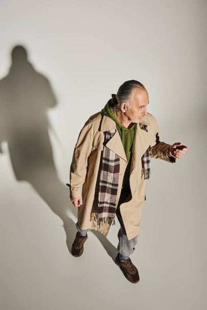 full length of senior man in stylish casual attire standing on grey background with shadow and looking away, green hoodie, beige trench coat, plaid scarf, fashionable aging concept, high angle view - Foto, Imagem