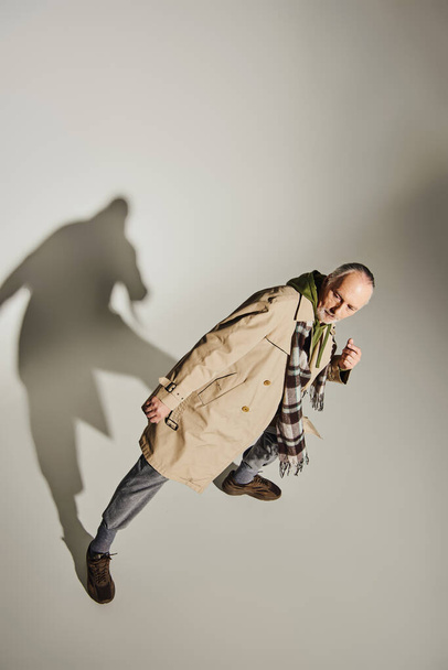 top view of confident senior model in plaid scarf, green hoodie and beige trench coat standing in stylish pose on grey background with shadow, fashionable aging concept - Φωτογραφία, εικόνα