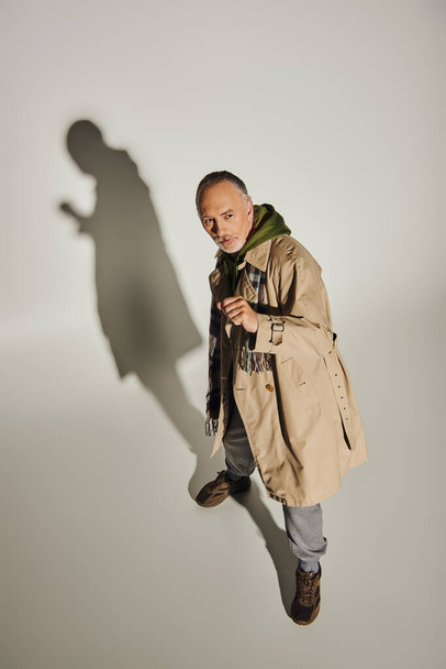 high angle view of expressive and stylish senior male model looking away on grey background with shadow, grey haired man in beige trench coat, plaid scarf and green hoodie, fashion and age concept - Φωτογραφία, εικόνα