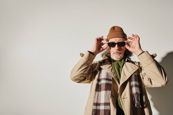 bearded senior man in beanie hat, plaid scarf and stylish trench coat looking at camera while standing on grey background, hipster style, expressive individuality, trendy aging concept - Foto, Imagen