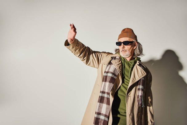modieuze senior man in donkere zonnebril, muts, beige trench jas en ruitvormige sjaal staande met uitgestrekte en weg te kijken op grijze achtergrond, hipster stijl, expressieve persoonlijkheid - Foto, afbeelding