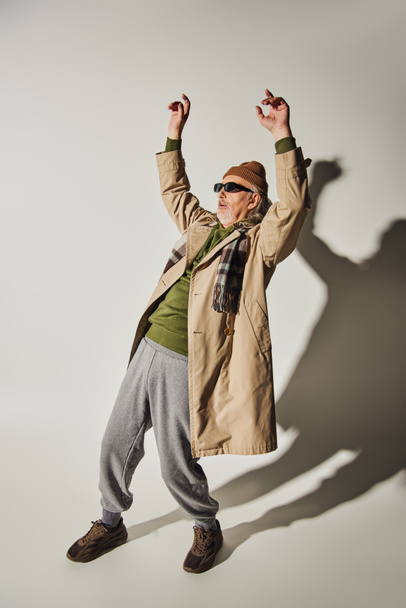 full length of expressive hipster style senior man in dark sunglasses, beanie hat, beige trench coat and plaid scarf posing with raised hands on grey background with shadows, dance - Photo, Image