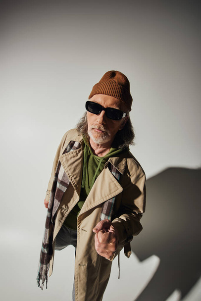 hipster style, trendy casual clothes, senior man in dark sunglasses, beanie hat and plaid scarf looking at camera on grey background with shadow, fashionable lifestyle and positive aging concept - Photo, Image