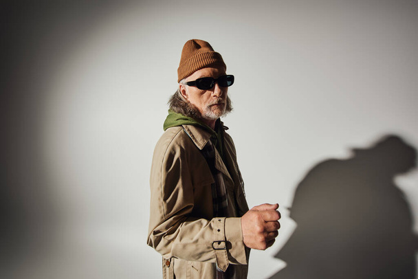 senior male model in dark sunglasses and stylish casual clothes looking at camera on grey background with shadow, beanie hat, beige trench coat, plaid scarf, expressive personality - Φωτογραφία, εικόνα