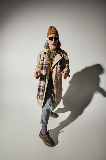 plná délka módní a chladný stárnoucí muž v tmavých slunečních brýlích, čepice a béžový trench kabát ukazuje prsty na kameru na šedém pozadí se stínem, hipster styl senior model - Fotografie, Obrázek