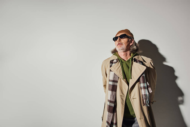 пожилой человек в стильной повседневной одежде и темных солнцезащитных очках стоя на сером фоне с тенью и глядя в сторону, хипстерский тренд, шапочка шапочка, бежевый плащ, мода и возрастная концепция - Фото, изображение