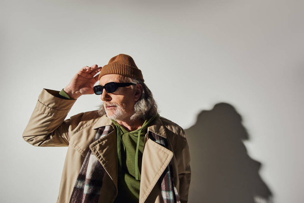 anciano hombre mayor de pelo gris y barbudo en gafas de sol oscuras, gabardina beige y bufanda a cuadros tocando sombrero de gorro y mirando hacia otro lado sobre fondo gris con sombra, estilo hipster, individualidad - Foto, imagen