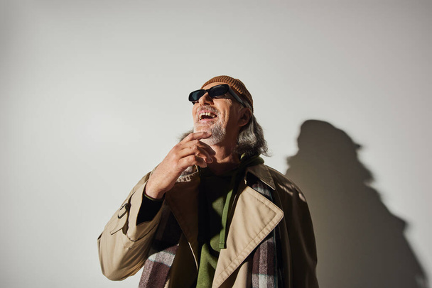 vrolijk en stijlvol verouderingsconcept, opgewonden hipster stijl senior man in donkere zonnebril, muts en beige trench jas die wegkijkt en lacht op een grijze achtergrond met schaduw - Foto, afbeelding