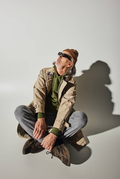 homme hipster senior en bonnet, lunettes de soleil sombres et trench coat beige assis avec jambes croisées et regardant loin sur fond gris avec ombre, concept de vieillissement à la mode - Photo, image