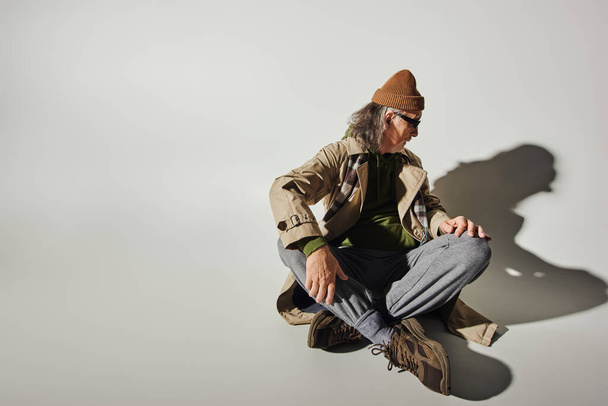 Mann im trendigen Hipster-Stil mit Hut, dunkler Sonnenbrille, beigem Trenchcoat und Turnschuhen, der seinen eigenen Schatten betrachtet, während er mit überkreuzten Beinen auf grauem Hintergrund mit Kopierraum sitzt - Foto, Bild