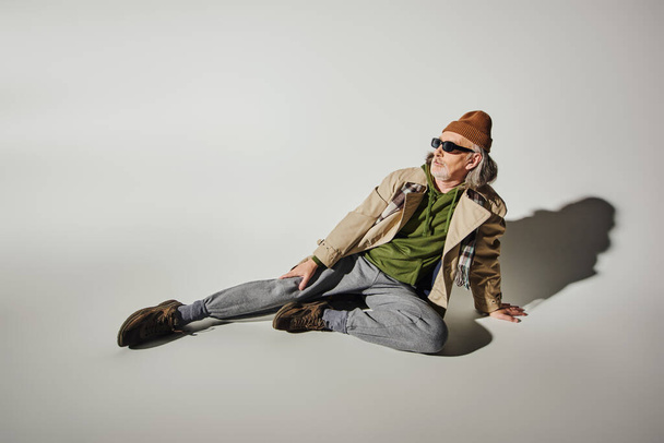 full length of fashionable senior male model sitting and looking away on grey background with shadow, dark sunglasses, beanie hat, beige trench coat, hipster style, positive aging concept - Photo, Image