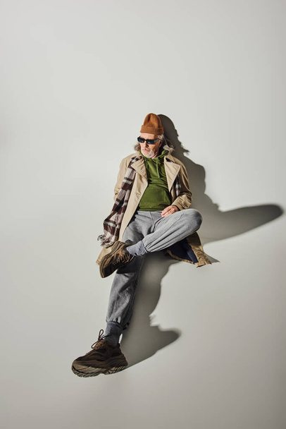 high angle view of senior male model in trendy hipster style attire and dark sunglasses laying and looking away on grey background with shadow, beanie hat, beige trench coat, fashion and age concept - Photo, Image