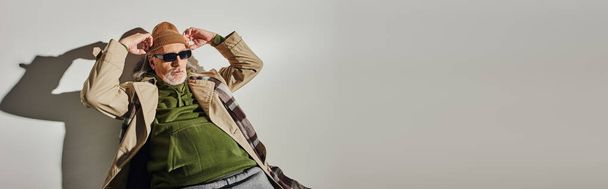 homem estilo moderno hipster em óculos escuros e casaco de trincheira bege ajustando chapéu gorro enquanto posando em fundo cinza com sombra, envelhecimento com conceito de estilo, banner - Foto, Imagem