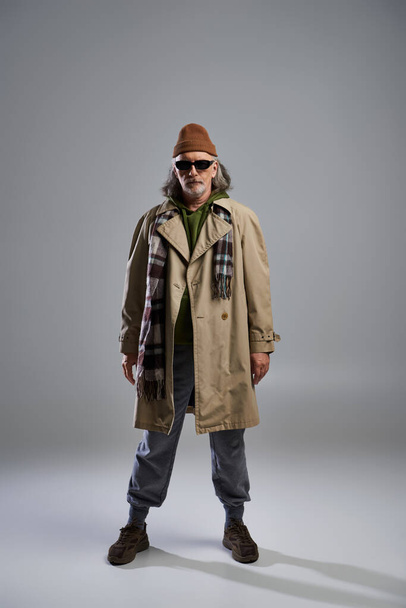 front view of trendy and bearded senior man in dark sunglasses, beanie hat, beige trench coat and sneakers standing on grey background, hipster fashion, trendy aging concept, full length - Photo, Image