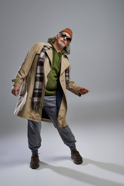 full length of aged senior model in hipster style outfit posing on grey background, beanie hat, dark sunglasses, beige trench coat, sneakers, fashionable lifestyle and positive aging concept - Photo, Image