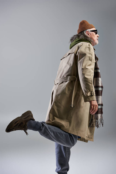 vista lateral do modelo masculino sênior em chapéu gorro, óculos escuros e casaco de trincheira bege posando em fundo cinza, pose elegante, moda hipster, conceito de envelhecimento na moda, tiro de moda - Foto, Imagem