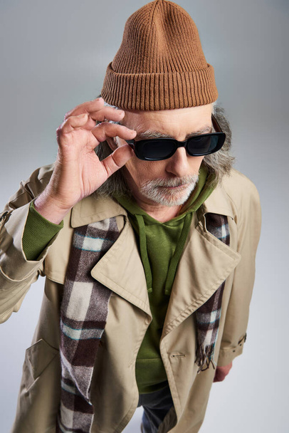 fashionable and bearded man adjusting dark sunglasses while standing on grey background, hipster fashion, beanie hat, beige trench coat, plaid scarf, aging with style concept, fashion shoot - Photo, Image