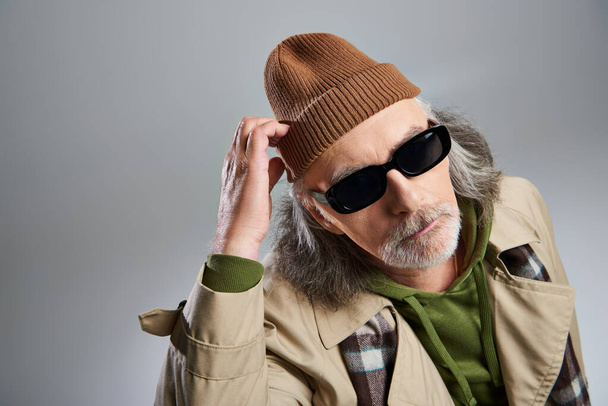 portrét přemýšlivého staršího muže v tmavých slunečních brýlích a béžovém plášti, dotýkající se čepice a hledící na kameru na šedém pozadí, hipsterská móda, stylový a pozitivní koncept stárnutí - Fotografie, Obrázek