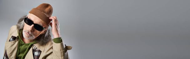 Porträt eines älteren männlichen Models mit dunkler Sonnenbrille und beigem Trenchcoat, dunkle Sonnenbrille einsetzend und in die Kamera blickend auf grauem Hintergrund, gealterter Hipster-Mann, Banner mit Kopierplatz - Foto, Bild