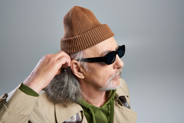 portrét staršího a vousatého hipsterského staršího muže v tmavých slunečních brýlích, čepici a béžovém plášti upravujícím šedé vlasy a odvracejícím se pohledem na šedé pozadí, módní stárnutí - Fotografie, Obrázek