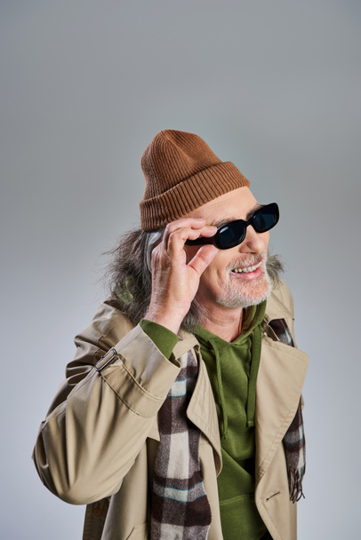retrato de hombre barbudo de estilo hipster sénior y alegre en gorro sombrero y gabardina beige ajustando gafas de sol oscuras, riendo y mirando hacia otro lado sobre fondo gris - Foto, imagen