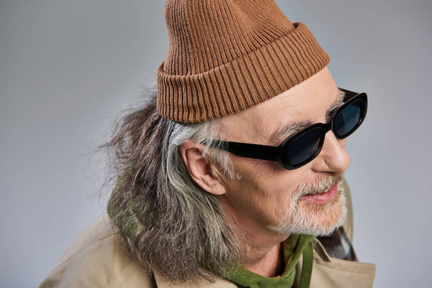 portrait of grey haired and cheerful senior man in fashionable hipster style outfit, beanie hat and dark sunglasses smiling and looking away on grey background, happy aging concept - Photo, Image