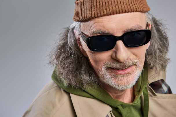 portrait d'un homme âgé souriant aux cheveux gris et à la barbe toilettée, portant des lunettes de soleil sombres, un bonnet et un imperméable regardant la caméra sur fond gris, une mode hipster, un concept de vieillissement heureux et tendance - Photo, image