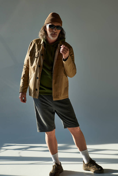 full length of aged bearded man in beanie hat, dark sunglasses, jacket and shorts looking at camera while standing on grey background with lighting, hipster fashion, trendy aging concept - Photo, Image