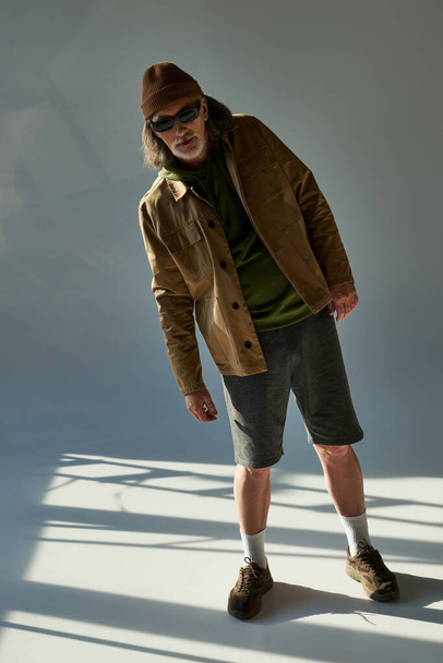 full length of senior male model looking at camera on grey background with lighting, aged hipster man in dark sunglasses, beanie hat, jacket and shorts, fashionable lifestyle concept - Foto, Imagem