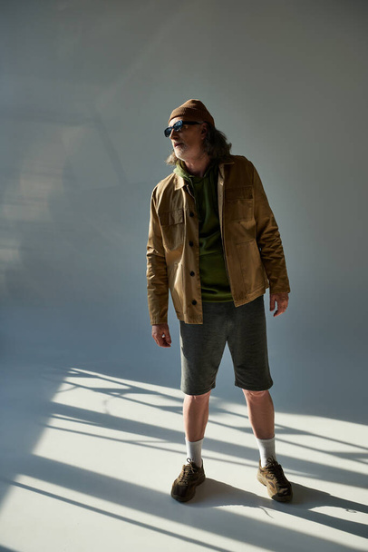 full length of senior and bearded man in dark sunglasses, beanie hat, jacket and shorts looking away while standing on grey background with lighting, hipster trend, expressive personality - Foto, imagen