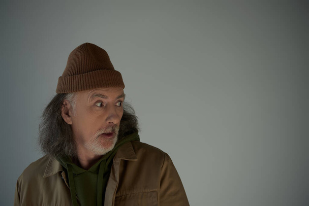 shocked hipster style man in beanie hat and brown jacket standing with open mouth and looking away on grey background, aging population lifestyle concept, copy space - Φωτογραφία, εικόνα