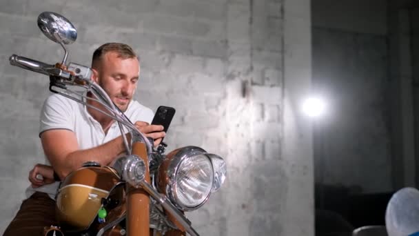 Ein Mann hält ein Smartphone in der Hand, während er auf einem Retro-Motorrad in einer Garage sitzt. Schönes Motorrad. - Filmmaterial, Video