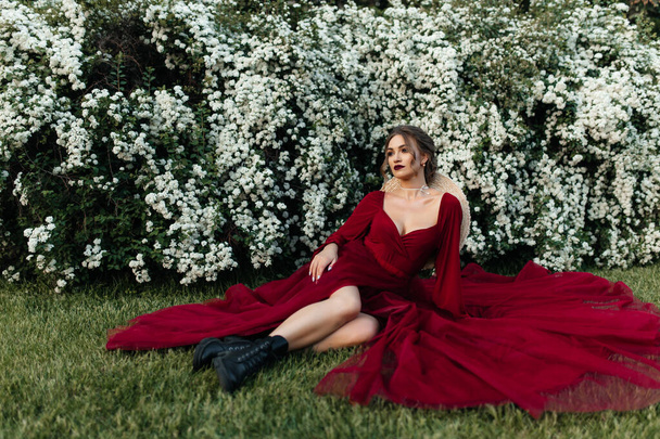 Kırmızı elbiseli güzel kız beyaz çiçekli çalıların yanında oturuyor. - Fotoğraf, Görsel