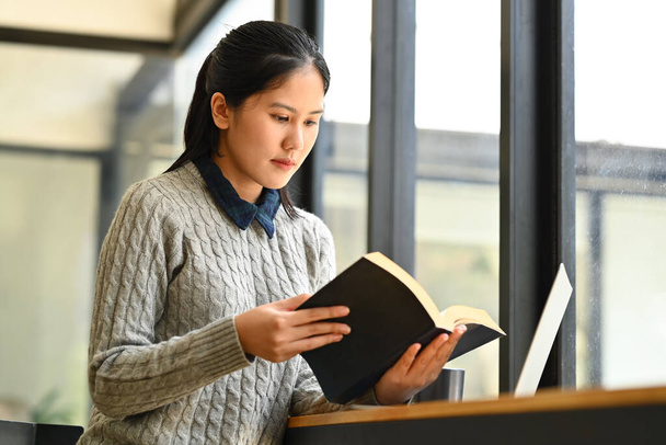 Békés ázsiai üzletasszony fontos jegyzeteket olvas a személyes napi tervezőben, laptopja előtt ül az irodában.. - Fotó, kép