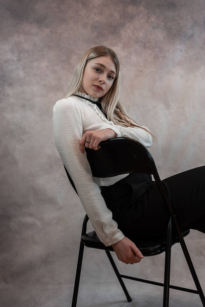Pretty kobieta model nosić białą bluzkę i czarne spodnie siedzi na krześle i patrząc na aparat fotograficzny, studio strzelać - Zdjęcie, obraz