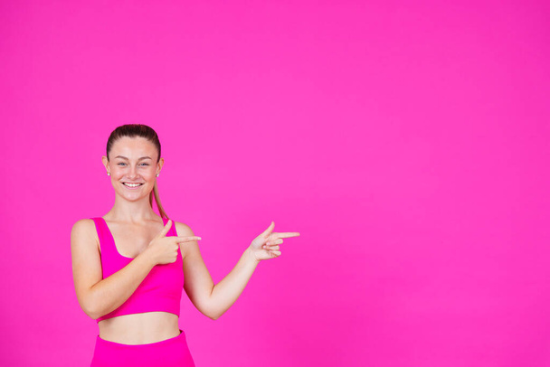 Smiling positive sportswoman pointing at message on pink colourful copy space. Young woman looking at camera and showing advertisement. - Foto, Bild
