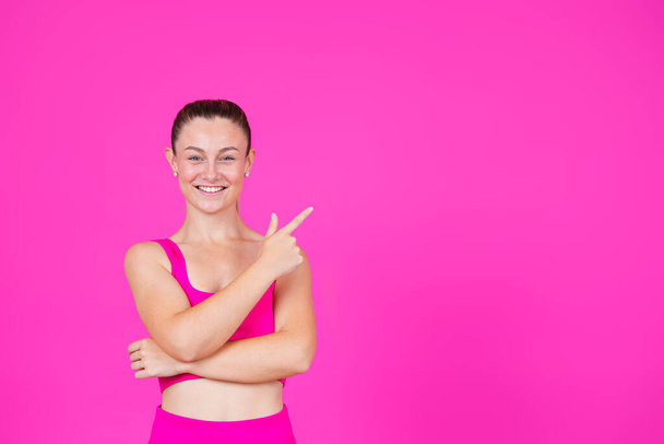 Спортивная позитивная женщина улыбается и смотрит в камеру. Женщина указывает на пространство для копирования, изолированное на розовом фоне. - Фото, изображение