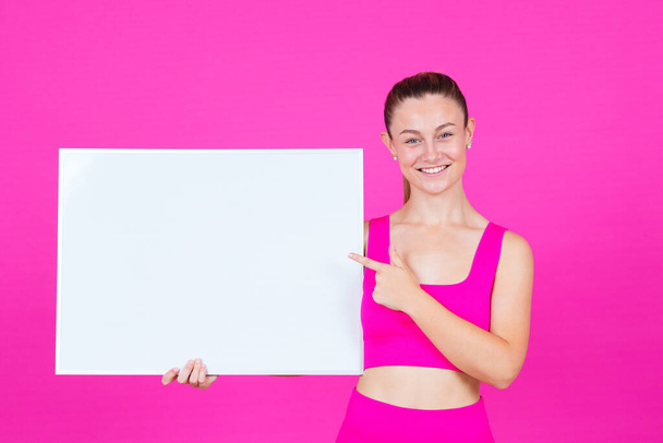 Smiling sportswoman showing empty billboard. Healthy instructor or coach pointing at advertisement board isolated on colourful background. - Photo, Image