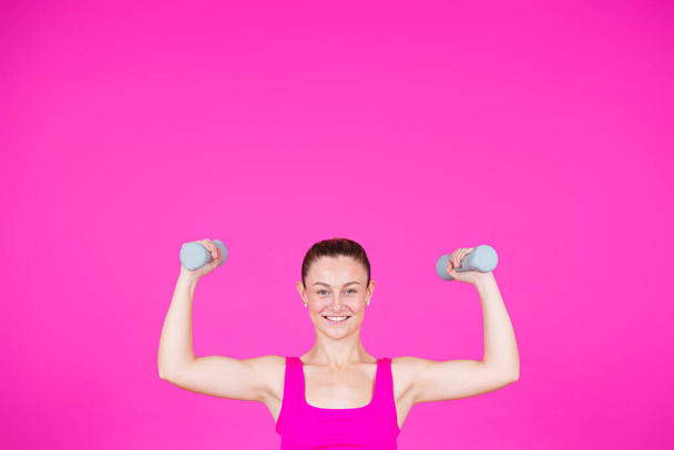 Lächelnde starke Sportlerin beim Heben von Gewichten isoliert auf rosa Hintergrund mit Kopierraum. junge Frau blickt in Kamera. - Foto, Bild