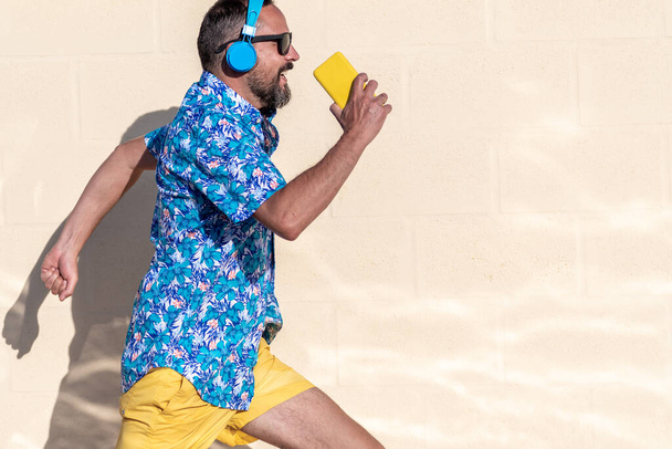 jovem homem hipster barbudo vestido com camisa havaiana, óculos de sol e fones de ouvido sem fio corre enquanto sorri. segura seu smartphone amarelo no espaço hand.copy. Fundo branco - Foto, Imagem