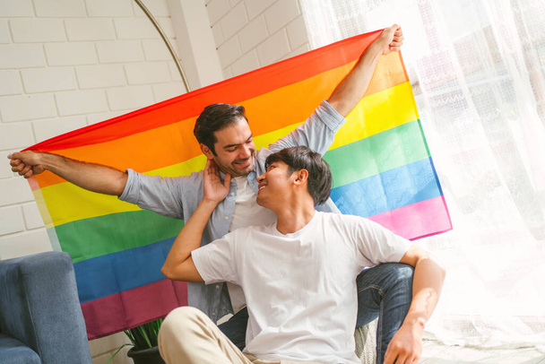 Щаслива пара кауказійських і азійських ЛГБТ-компаній, які сидять на дивані і розмахують веселковим прапором ЛГБТ Гордості разом у вітальні вдома. Різноманітність ЛГБТ-відносин. Концепція пари геїв. - Фото, зображення
