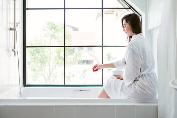 Beautiful happy Caucasian woman in a stylish bathrobe sitting on the tub with rose petals in her hand. Woman in bathrobe waiting for filling water in bathtub and spread rose petals before soaking. - Foto, Imagem